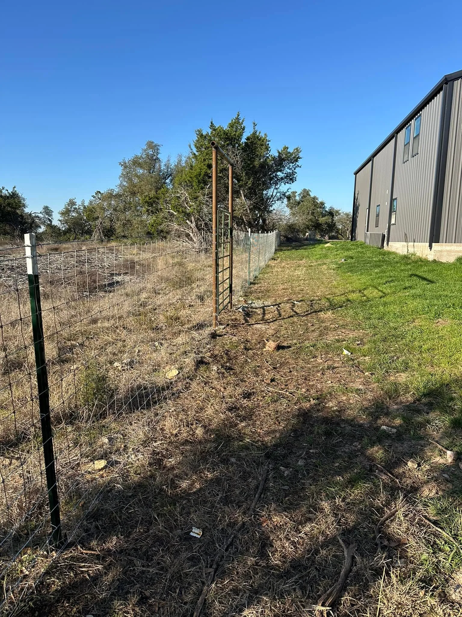 Green t post woven wire fence pipe gate frame beside gray metal building